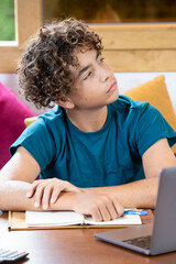 Portrait of a thoughtful teenage boy taking a break while studying at home with a laptop and notebook. Student looking away with a reflective, curious expression during an online lesson © NataSao