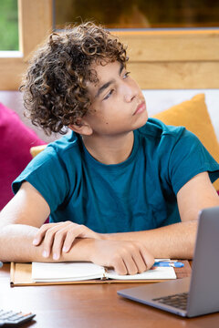 Portrait of a thoughtful teenage boy taking a break while studying at home with a laptop and notebook. Student looking away with a reflective, curious expression during an online lesson