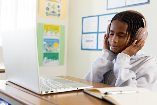 African American teen in hoodie sitting at desk with pink headset using laptop notebook, copy space