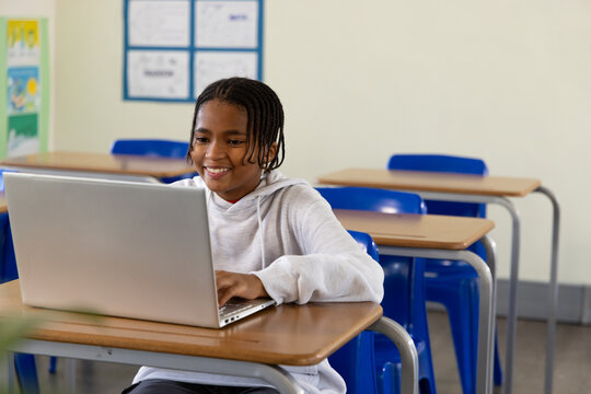 African male youth student sitting at front classroom wooden desk typing on silver laptop, smiling
