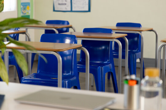 Blue plastic student chairs are lining classroom rows with wood-top desks, laptop, markers, plant