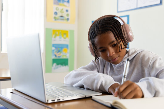 African American youth in hoodie and pink headset writing notebook at desk in class, open laptop