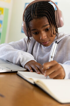 African American teen male studying at table in gray hoodie, pink headset, writing notebook, laptop