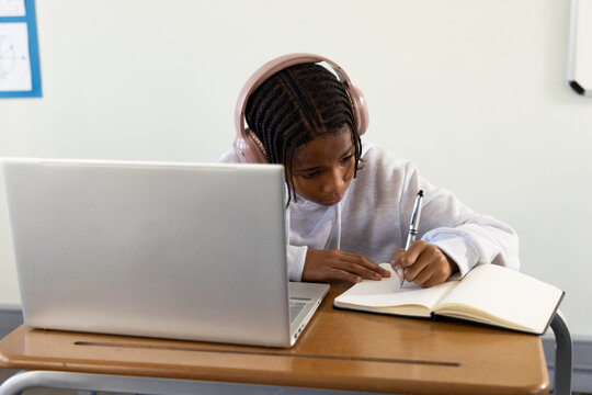 African American teen writing in notes using laptop at school desk wearing pink headset and hoodie