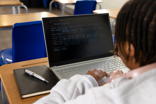 African American teenage male coding on silver laptop at school desk with notebook, pink headphones