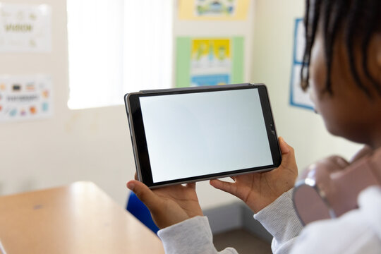 Teenage male holding tablet horizontally at eye level in classroom, wearing over-ear headphones