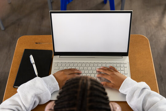 African American teenage male typing on laptop at school desk with black notebook, wearing sweater