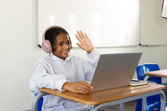 African American teen sitting in class at desk waving at silver laptop with pink headset