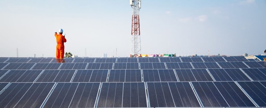 Worker stands tall on solar panels under clear blue skies. Orange suit contrasts with dark panels and distant tower. Sunlight reflects off rows of modern energy collectors