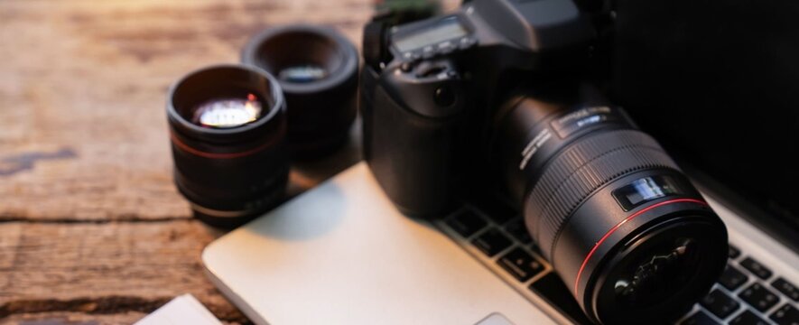 Camera rests beside laptop on rustic wood surface. Lenses stand nearby, ready for creative use. Professional gear suggests photography projects. Setup implies planning or editing for digital media