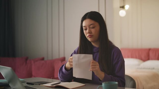 Young Asian woman opening envelope and reading bank statement letter at home