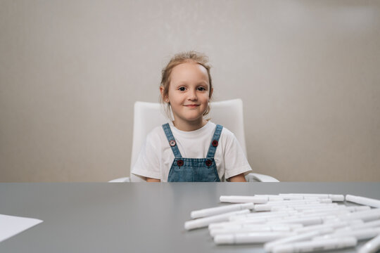Charming portrait of young girl sitting at grey table. Child wears denim overalls and smiling looking at camera. Minimalist background with soft lighting. Concept for childhood, creativity, education.