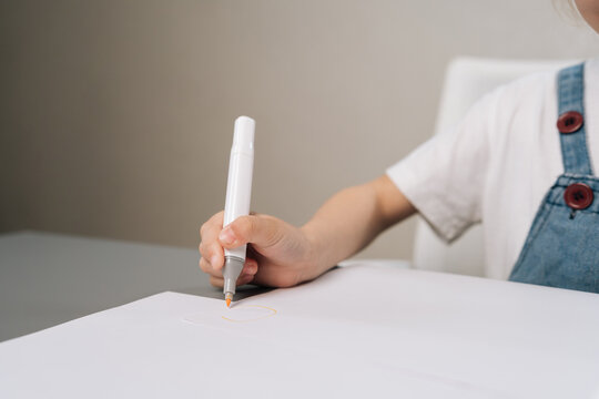 Close-up shot of young child hand holding white marker, drawing orange circle on paper. Scene has soft lighting and neutral colors, evoking creativity, focus and learning.