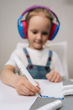 Studio portrait of young girl engaging in creative drawing activity with blue marker pen on white paper, wearing noise cancelling headphones for focused learning and artistic expression.
