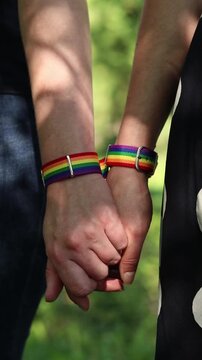 Two people holding hands with LGBTQ+ rainbow bracelets. Close-up of hands holding together with pride symbols. Same-sex couple holding hands on blurred natural background. Love and equality concept