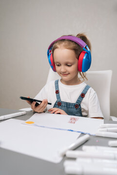 Studio portrait of young girl in bright headphones at desk, drawing with markers and looking at smartphone. Modern childhood concept of e-learning, creativity, digital education. Calm, focused mood.