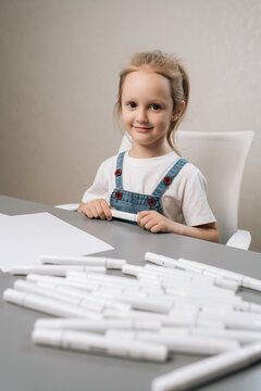 Vertical portrait of young girl with cheerful expression sitting at table with blank paper and numerous markers, developing creativity and artistic skills through drawing.