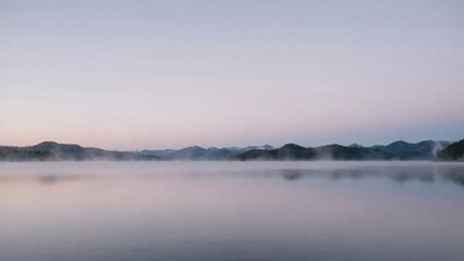 Fototapeta premium Serene morning mist over calm mountain lake waters during early dawn twilight hour with framed distant forested mountains under soft pastel sky