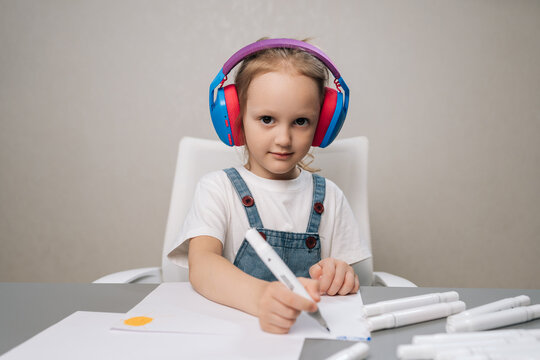 Focused young girl in vibrant headphones draws with markers at table. Child looking directly at camera, concentrating on creative activity. Concept for homeschooling, art, childhood development.