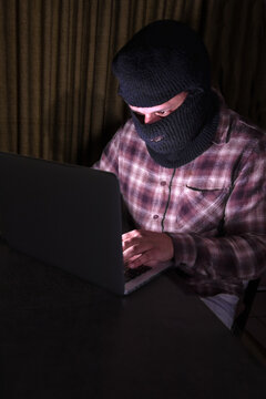 Cybercriminal wearing a black balaclava typing on a laptop in a dark room