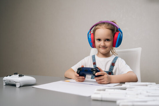 Studio portrait of young girl enjoying leisure time, wearing vibrant headphones and holding dark controller, playing video game at table with drawing and markers.