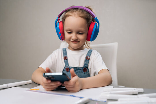 Happy child at white desk wears bright headphones watching mobile phone. Concept of modern childhood, homeschooling, screen time and digital entertainment. Soft light, neutral background.