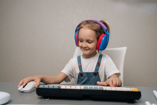 Smiling young child student in headphones enjoys e-learning or gaming. Focused child using keyboard and mouse for online education or entertainment at home. Modern technology for kids development.