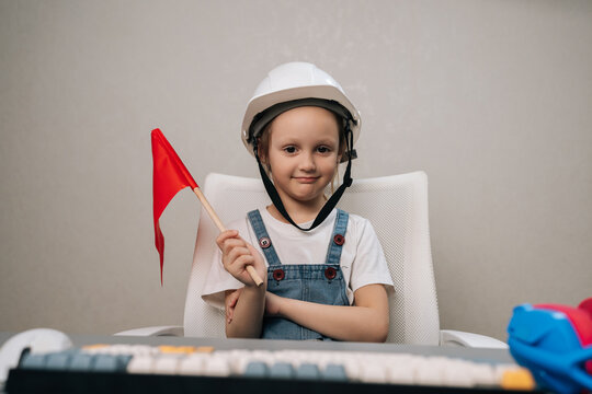 Young girl in denim overalls and white construction helmet sitting at desk holding small red flag, smiling confidently as she pretends to be an engineer, dreaming of future.