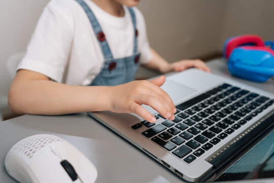 Close-up shot of young childs hands typing on laptop keyboard. Kid engaged in e-learning or homeschooling. Modern home setting with mouse and headphones nearby. Concept of digital literacy.