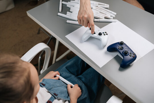 Child girl hand pointing at game controller buttons, teaching young child holding marker about gaming and digital entertainment next to stack of markers.