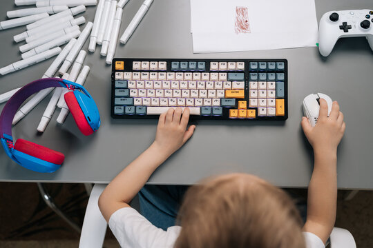 Child hands using typing colorful mechanical keyboard and white mouse, with headset, drawing game controller on grey desk, showing blend of learning and leisure activities.
