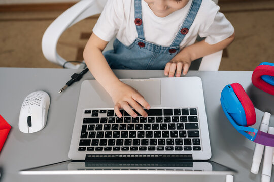 Top view of young boy in overalls studying on laptop. Child hand on trackpad showing focus on e-learning gaming. Bright, modern setup with headphones mouse. Homeschooling digital literacy concept.