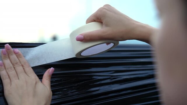 Woman attaching garbage bag with adhesive masking tape to broken car window. Driver fixing temporary patch on damaged automobile after accident or vandalism, close-up, slow motion.