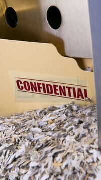 Vertical stock photo, manila folder labeled CONFIDENTIAL sitting dangerously close to an active paper shredder