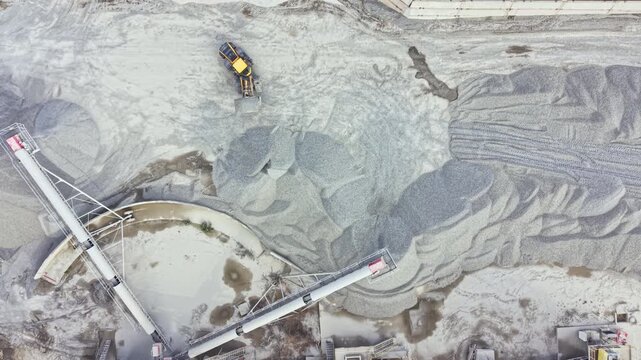 Heavy machines work in a granite quarry moving and processing stone material. The scene shows piles of gravel and machinery in action during daylight hours.