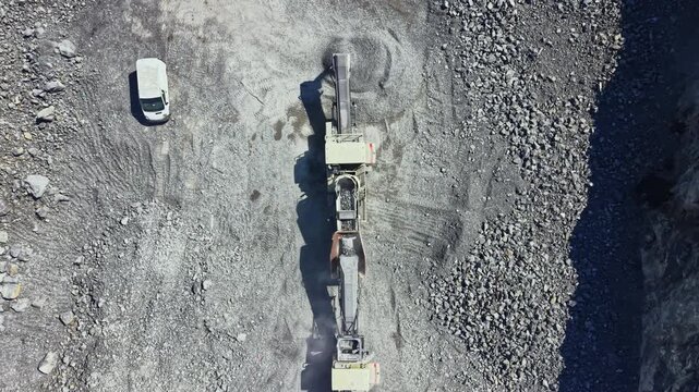 Heavy machinery is working in a granite quarry to cut and process rock. Workers are present while a truck is parked on the side. Dust is rising from the equipment as it operates.