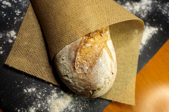 A loaf of bread wrapped in burlap cloth sits against a dark background with scattered flour, showing its golden crust peeking through