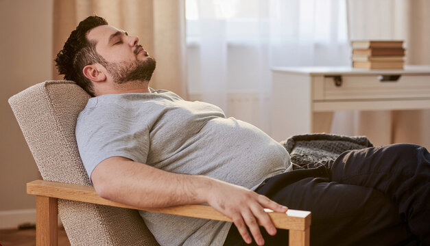 A relaxed man sleeping peacefully on a wooden armchair during a lazy day at home, sitting comfortably with his eyes closed in a bright living room, enjoying a quiet nap and deep relaxation.