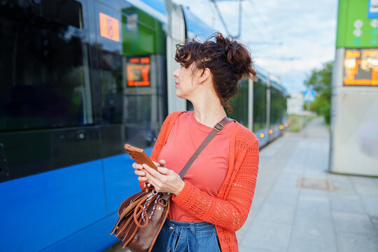 Alert young woman checking arriving tram while holding smartphone at station