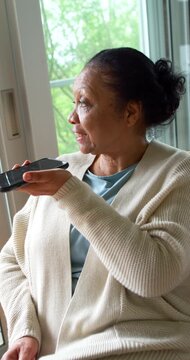 Vertical video: Senior African American woman using smartphone speaker in armchair by sliding door