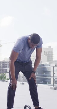 Vertical video: Man reaching and gripping 5kg kettlebell on roof in blue tee, gasping after set