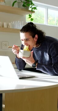 Vertical video: African American man eating noodles with bowl while working on laptop in kitchen