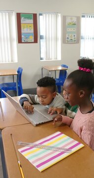 Vertical video: Diverse children leaning as boy pressing keys on laptop, learning at school desk