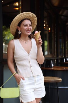 Smiling woman in hat and bag holding waffle cone with delicious ice cream at outdoor cafe