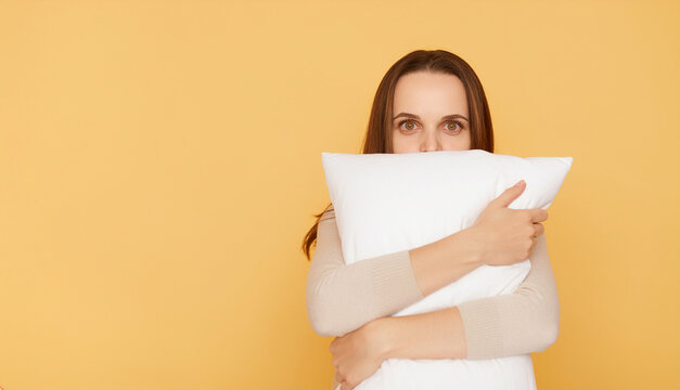 A young woman with brown hair hiding half of her face behind a white pillow while looking at the camera, standing in front of a solid pastel yellow background during a cozy lazy day.