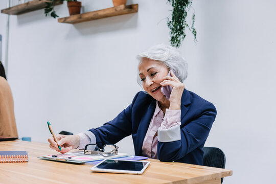 hispanic business mature woman talking and using phone in call at office. Latina senior or middle age female at workplace holding mobile phone