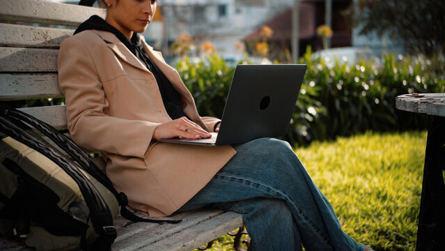 Young Hispanic woman using her laptop outdoors in a green space, representing remote work, productivity, technology, and a balanced modern lifestyle.