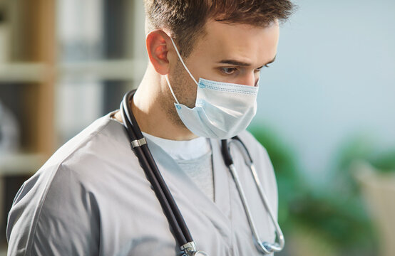 Close up portrait of pensive man doctor or nurse in blue face mask standing in medic office. Young male physician wearing stethoscope in clinic with copy space. Health care and medicine concept.