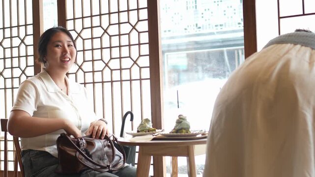 Woman smiles at table with tea and small green birds. Another person stands nearby, back turned. Traditional wooden window frames the scene. Good for cafes, cultural events, or relaxed dining scenes