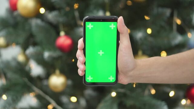 A blurred hand displays a phone with a green chroma key screen near a Christmas tree adorned with baubles and fairy lights. The image captures a modern holiday celebration atmosphere